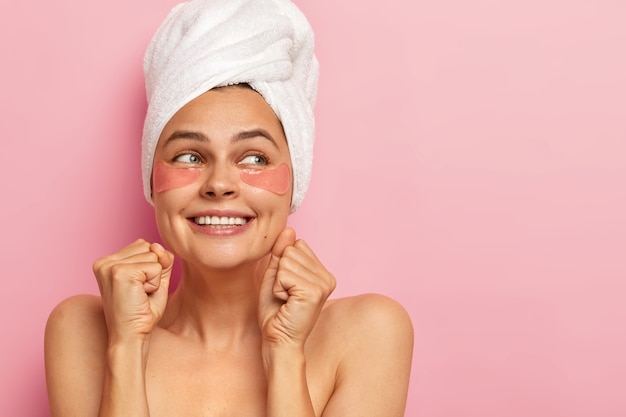 Woman clenches teeth and fists, looks away with glad expression, wears white soft towel on head, has naked shoulders