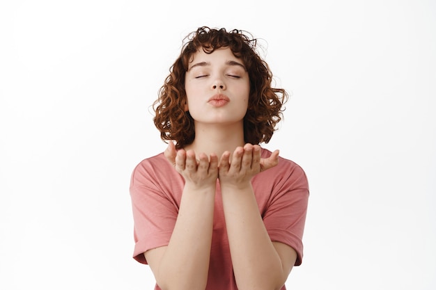 Foto gratuita la donna chiude gli occhi, manda un bacio d'aria, soffia mwah baciando con le mani aperte vicino alle labbra, in piedi in maglietta su bianco