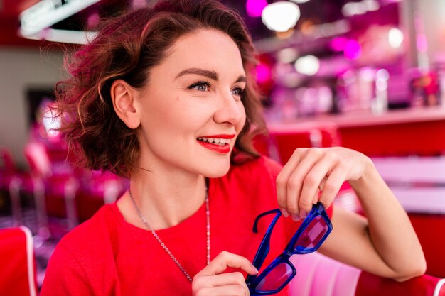 woman in colorful outfit in retro vintage 50's cafe sitting at table wearing red shirt, blue sunglasses having fun in cheerful mood, red lipstick make-up