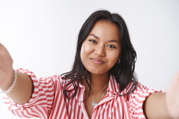 Foto gratuita la donna si avvicina per abbracciarci ritratto di amichevole adorabile e tenera fidanzata vietnamita che estende le mani tenendo la fotocamera inclinando la testa e sorridendo mentre guarda il fidanzato romantico