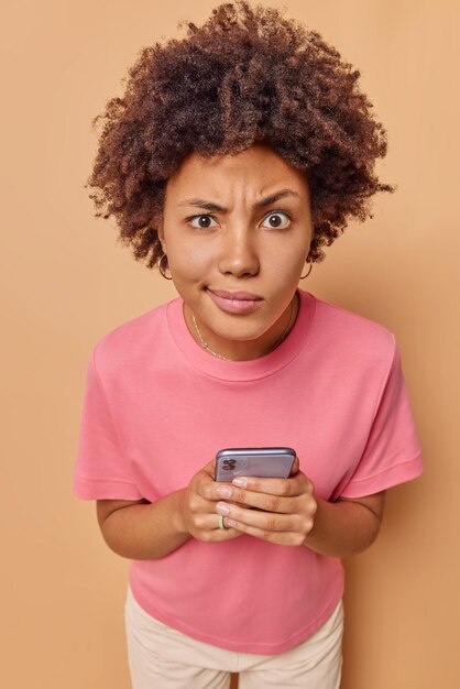 woman concentrated attentively at camera holds mobile phone sends text messages wears casual pink t shirt isolated on beige chats online in social network