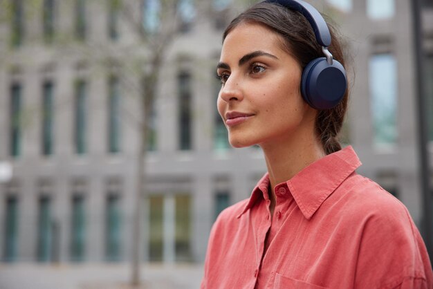 woman concentrated forward listens audio song wears red shirt has thoughtful face expression poses outdoor with blank copy space for your advertising content