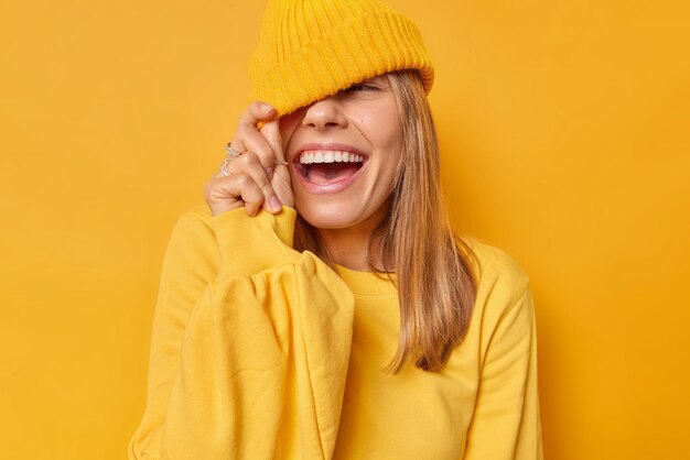 woman cons eyes with hat has joyful expression dressed in casual jumper isolated on bright yellow. Carefree teenage girl foolishes around has funny expression