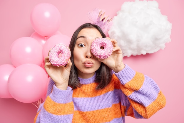 женщина против глаз с вкусными пончиками держит губы сложенными, одетая в повседневный джемпер, празднует день рождения, наслаждается праздником, изолирована на розовом