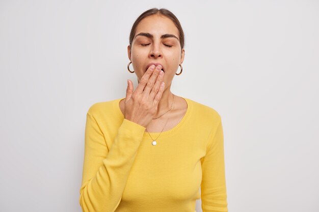 женщина против рот рукой держит глаза закрытыми, носит повседневный желтый свитер, чувствует усталость или сонный стоит на белом