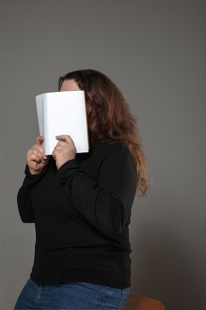 woman covering face with book while reading on grey wall