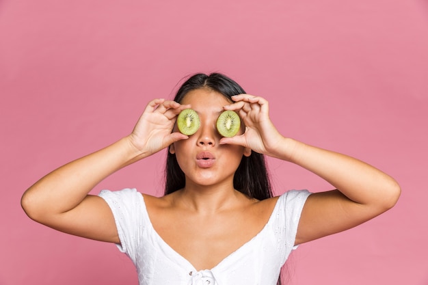 Woman covering her eyes with kiwi on pink surface