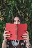 Foto gratuita donna che copre la bocca con il libro in piedi contro la pianta crescente