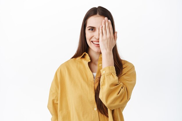 Woman covers one half of face and smiles at camera, before after concept, having nude natural makeup on, standing in yellow stylish oversized shirt, white background