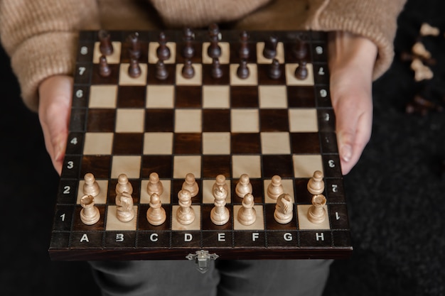 Free photo a woman in a cozy sweater holds a chessboard with wooden chess pieces set apart.