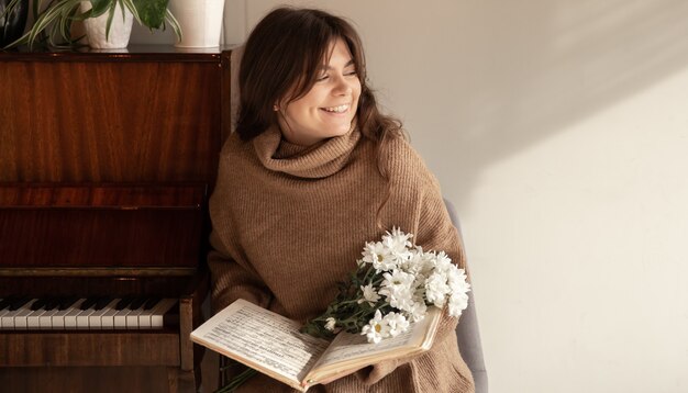 A woman in a cozy sweater holds a collection of notes and a bouquet of flowers, sitting near the piano, copy space.