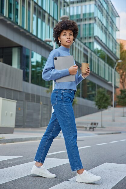 woman crosses road on crosswalk carries digital tablet and paper cup of coffee wears casual jumper jeans strolls in downtown returns after work