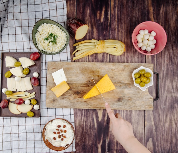 Foto gratuita una donna taglia formaggio olandese su un tagliere di legno con olive in salamoia e vari tipi di formaggio in vista dall'alto rustico