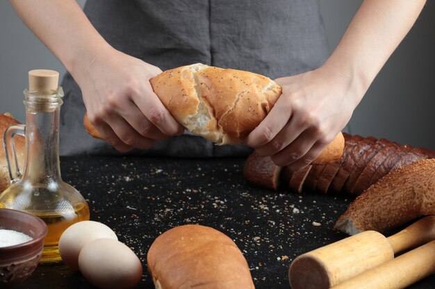 Woman cutting bread into half on dark table with eggs, flour bowl and glass of oil.