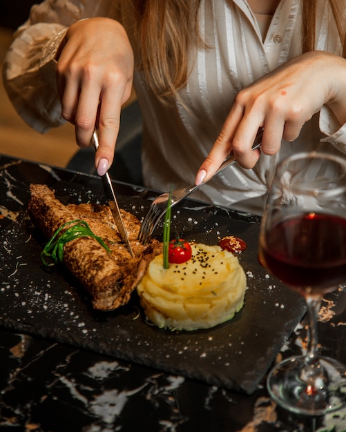 Женщина резки мяса стейк с картофелем в масках и стакан красного вина.