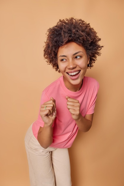 woman dances and shakes fists moves with rhythm of music has upbeat mood wears casual pink t shirt trouses looks away happily isolated on beige