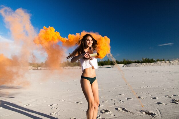 Woman dances with orange smoke on white beach under blue sky