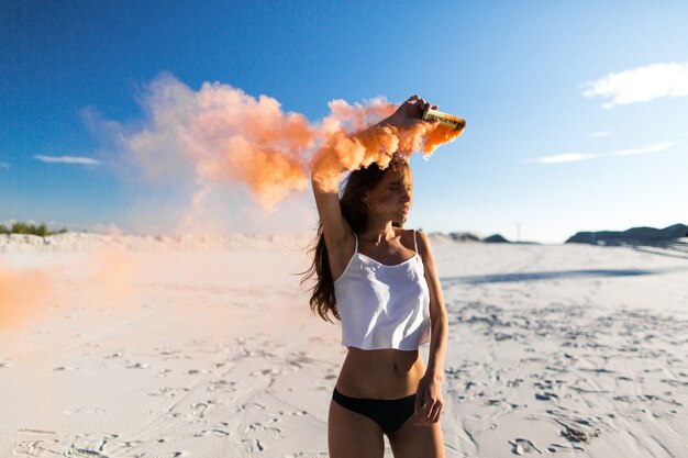 Woman dances with orange smoke on white beach under blue sky