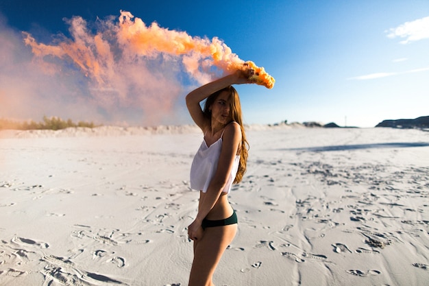 Woman dances with orange smoke on white beach under blue sky