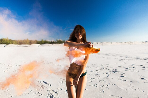 Woman dances with orange smoke on white beach under blue sky