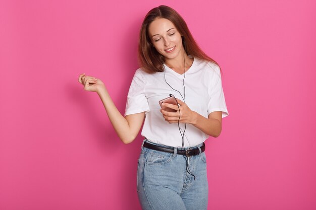 Женщина танцует с наушниками, слушая музыку через смартфон. Игривая счастливая улыбающаяся молодая, привлекательная леди, проводящая свободное время