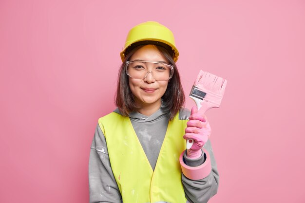 женщина-декоратор держит малярную кисть, защитные прозрачные очки, каску ремонтирует стены в квартире
