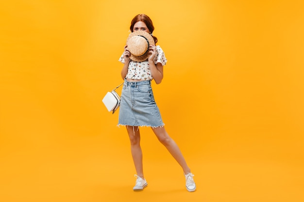 Woman in denim skirt and white cropped top covers her face with hat and looks into camera on orange background.