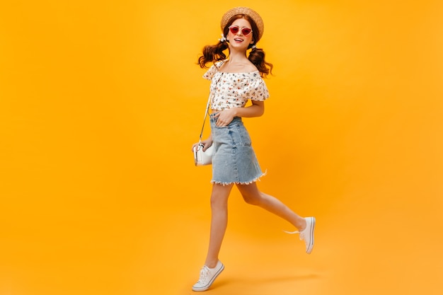 woman in denim skirt, white T-shirt and boater jumping on orange background. Woman in sunglasses smiling.