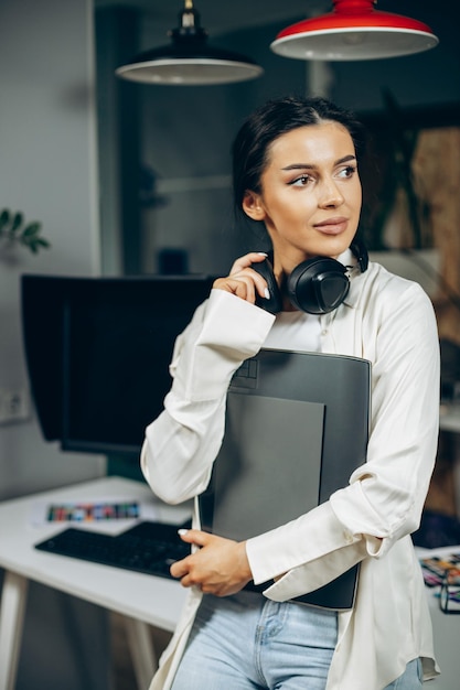 Woman designer with musical earphones standing in office holding folder