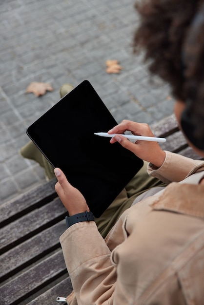 女性デザイナーがデジタルタブレットのダウンロードに取り組んでいますアプリケーションは、木製のベンチで屋外のスタイラスポーズで描画します