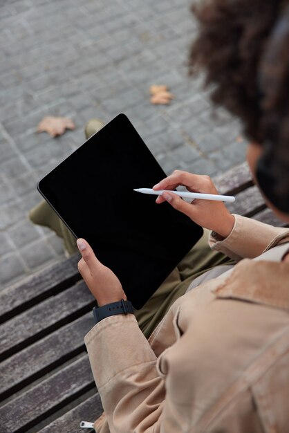 여성 디자이너는 나무 벤치에서 야외에서 스타일러스 포즈로 디지털 태블릿 다운로드 응용 프로그램을 그리는 작업을 합니다.