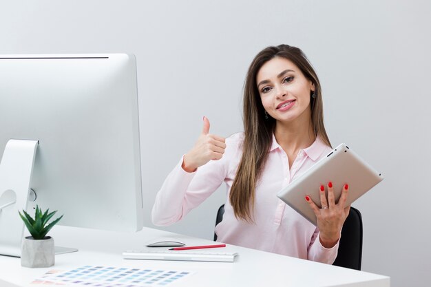 Woman at desk giving thumbs up while holding tablet