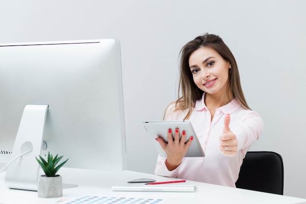 Woman at desk holding tablet and giving thumbs up