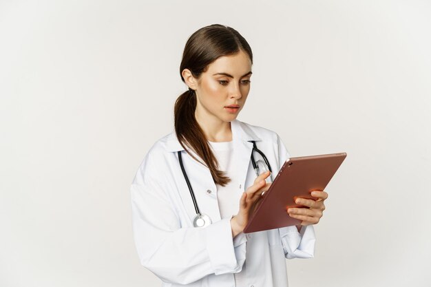 Woman doctor looking concerned at digital tablet reading with worried face expression wearing white ...