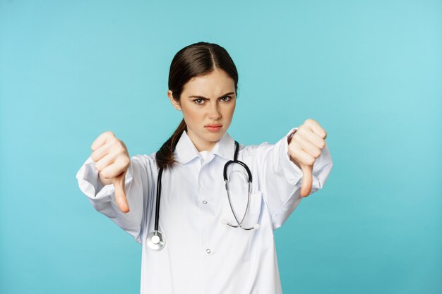 Женщина-врач-медицинский работник показывает большие пальцы вниз и хмурится, злится и не одобряет что-то стоящее в...