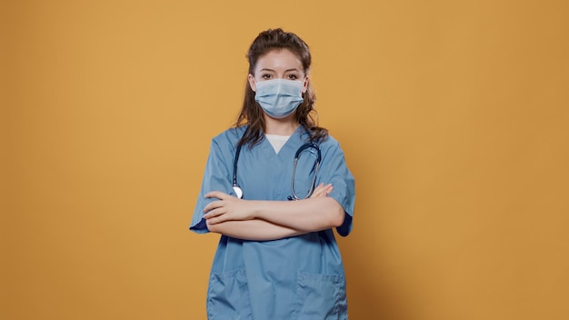 Woman doctor putting on covid mask for protection and posing confindent with arms crossed in studio with copy space background. Portrait of caucasian medic wearing protective uniform and stethoscope.