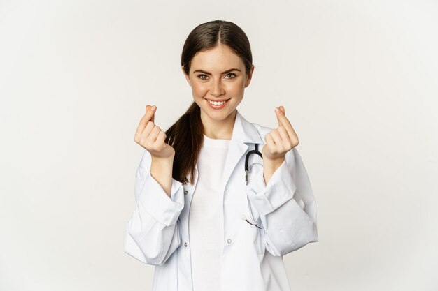 흰색 배경 위에 제복을 입고 조심스럽게 서서 웃고 있는 손가락 하트를 보여주는 여성 의사