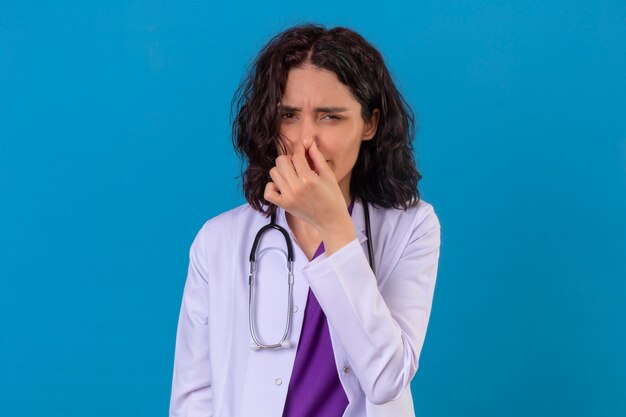 woman doctor wearing white coat with stethoscope closing nose with fingers feels unpleasant scent suffers from stench on isolated blue