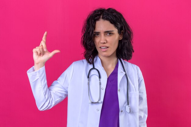 woman doctor wearing white coat with stethoscope gesturing with hands showing small size sign measure symbol on isolated pink