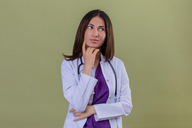 woman doctor wearing white coat and with stethoscope holding hand under her chin having doubts on isolated green