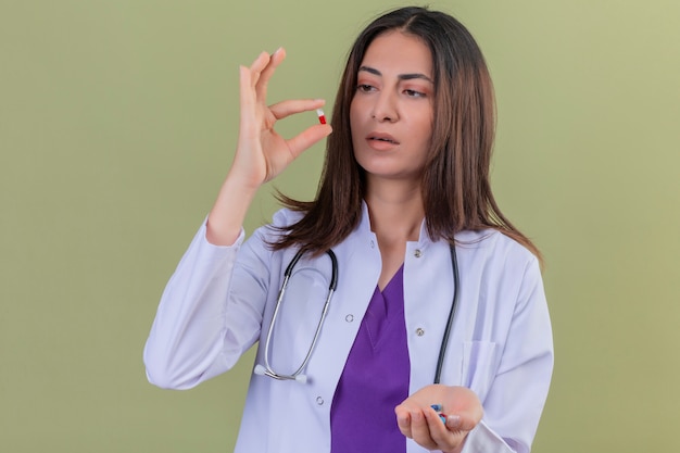 Donna medico che indossa camice bianco e con lo stetoscopio tenendo la pillola con le dita guardandolo con la faccia seria in piedi sul verde