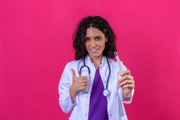 격리 된 분홍색에 서 행복 한 얼굴로 엄지 손가락을 보여주는 손에 온도계를 들고 청진기와 흰색 코트를 입고 여자 의사