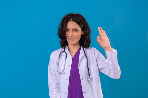 женщина-врач в белом халате со стетоскопом выглядит счастливой, улыбается, делает хорошо, знак, стоящий на изолированном синем