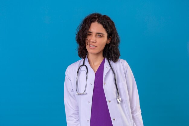 женщина-врач в белом халате со стетоскопом выглядит радостной, улыбаясь и подмигивая, стоя на изолированном синем