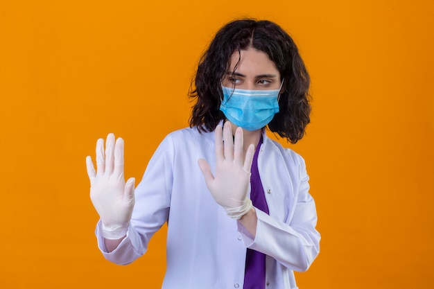 woman doctor wearing white coat with stethoscope in medical protective mask holding her hands up telling do not come closer on isolated orange