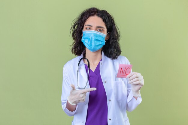 woman doctor wearing white coat with stethoscope in medical protective mask holding reminder paper with no word pointing with finger to it on isolated green