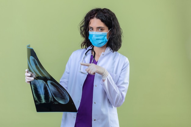 Foto gratuita donna medico che indossa camice bianco con uno stetoscopio in maschera protettiva medica tenendo i raggi x dei polmoni puntando ad esso con il dito con faccia seria sul verde isolato