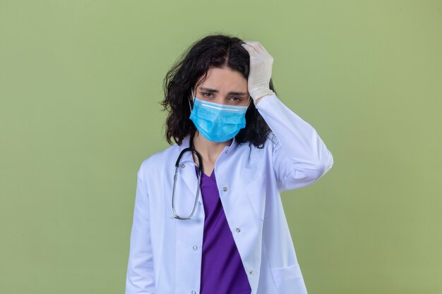 woman doctor wearing white coat with stethoscope in medical protective mask looking tired and onworked standing with hand on head having headache on isolated green