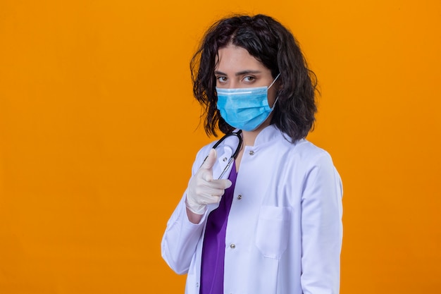 woman doctor wearing white coat with stethoscope in medical protective mask pointing at camera with finger with serious face standing on isolated orange
