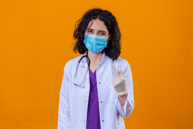 woman doctor wearing white coat with stethoscope in medical protective mask showing middle finger smiling cheerfully standing on isolated orange